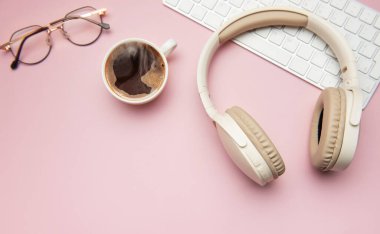 Kablosuz kulaklıklar, klavye ve gözlükler pembe arka planda bir fincan kahvenin yanında, ev dekorasyonundan rahatlatıcı bir çalışmayı temsil ediyor.