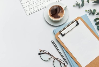 Beyaz arkaplanda klavye, kahve, dosya, bardak ve okaliptüs dalları olan ofis masası