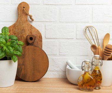 Tahta kesme tahtaları, taze fesleğen, havan, çırpma, baharatlar ve yağ şişeleri ahşap tezgahın üstünde beyaz tuğla duvara karşı dizilerek rahat bir mutfak ortamı yaratıyor.