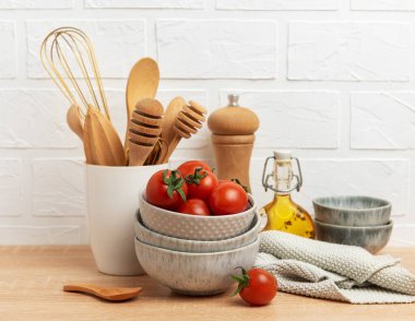 Tahta kaşıklar, çırpılar, kaseler, taze domatesler, zeytinyağı, biber değirmeni ve bulaşık bezi ahşap bir masaya dizilmiş beyaz tuğla bir duvara karşı.