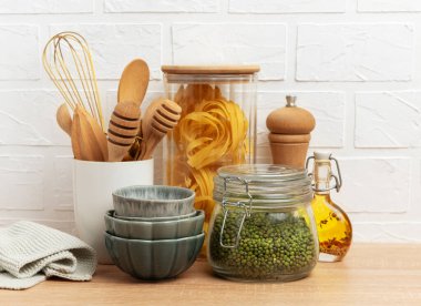 Ahşap mutfak gereçleri, makarna, mung fasulyesi, yağ ve kaseler beyaz tuğla duvara karşı ahşap bir tezgahta dizilmiş, rahat bir mutfak sahnesi yaratıyor.