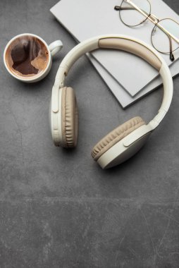 Bej kablosuz kulaklık, buharlı kahve fincanı, gözlük ve not defterleri gri masa üzerinde modern çalışma alanı şeklinde duruyor.