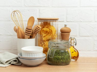 Ahşap mutfak gereçleri, makarna, mung fasulyesi, yağ ve kaseler ahşap bir tezgahın üzerinde beyaz tuğla duvara karşı dizilmiş rahat bir mutfak sahnesi oluşturur.