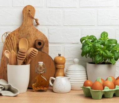 Ahşap mutfak aletleri, taze yumurtalar, fesleğen bitkisi ve zeytinyağı ahşap bir tezgahın üzerinde beyaz tuğla bir duvara karşı rahat bir atmosfer yaratıyor.