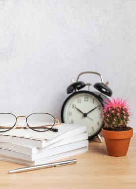 Gözlükler bir kalem, alarm saati ve küçük kaktüs bitkisinin yanında bir yığın defterin üzerinde oturur ve sakin bir çalışma alanı ortamı yaratır.