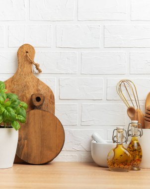 Tahta kesme tahtaları, taze fesleğen, havan, çırpma, baharatlar ve yağ şişeleri ahşap tezgahın üstünde beyaz tuğla duvara karşı dizilerek rahat bir mutfak ortamı yaratıyor.