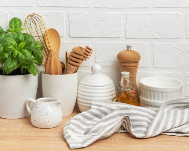 Çeşitli mutfak gereçleri, fesleğen bitkisi ve beyaz tuğla bir duvara karşı ahşap bir masaya dizilmiş keten bir peçete rahat ve davetkar bir atmosfer yaratıyor.