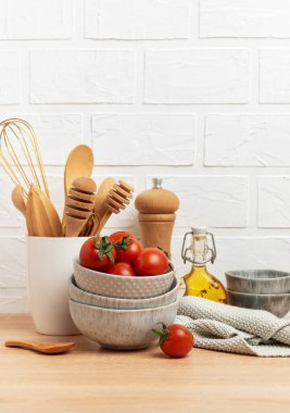 Tahta kaşıklar, çırpılar, kaseler, taze domatesler, zeytinyağı, biber değirmeni ve bulaşık bezi ahşap bir masaya dizilmiş beyaz tuğla bir duvara karşı.