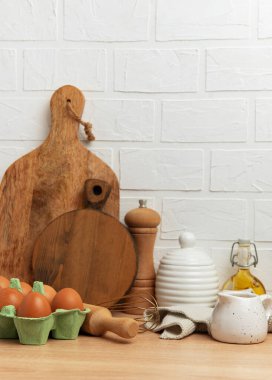 Tahta kesme tahtaları, yumurtalar, yağ, biber değirmeni, çırpma makinesi, merdane ve seramik masa takımı ahşap bir masada beyaz tuğla bir duvara karşı samimi bir mutfak sahnesi oluşturur.