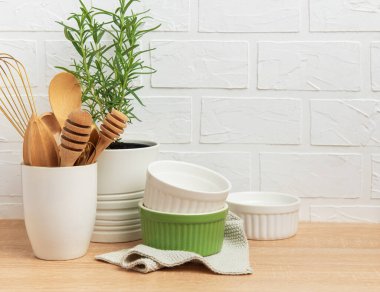 Ahşap mutfak gereçleri, biberiye bitkisi ve beyaz tuğla duvara karşı ahşap bir tezgahta dizilmiş seramik ramekinler, samimi bir mutfak sahnesi hazırlıyorlar.