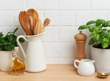 Ahşap mutfak gereçleri, taze fesleğenler, baharatlar ve yağlar beyaz tuğla bir duvara sergilenmiş, sıcacık bir mutfak ortamını somutlaştırıyor.