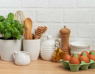 Mutfak tezgahında taze yumurtalar, fesleğen bitkisi, yağ, baharatlar ve beyaz tuğla bir duvara dayanan çeşitli mutfak aletleri ev yapımı bir atmosferi somutlaştırıyor.