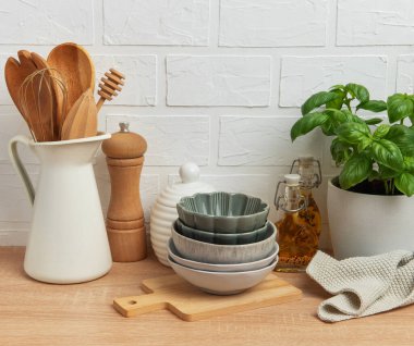 Kâseler, tahta kaşıklar, bir biber değirmeni, yağ şişeleri ve fesleğen bitkisi de dahil olmak üzere çeşitli mutfak malzemeleri beyaz tuğla bir duvara karşı ahşap bir tezgahta güzelce düzenlenmiştir.