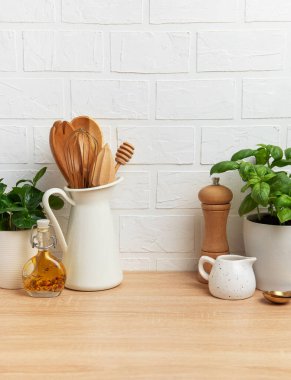Ahşap mutfak gereçleri, taze fesleğen, yağ ve baharatlar beyaz tuğla duvara karşı ahşap bir tezgahta dizilmiş, rahat bir mutfak sahnesi yaratıyor.