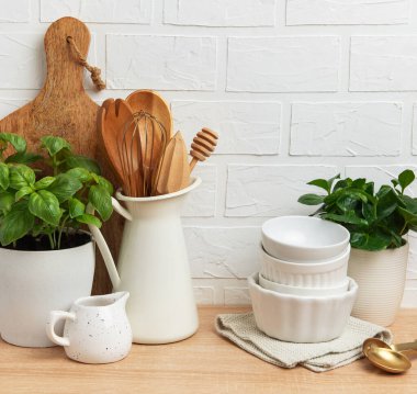 Ahşap mutfak aletleri, seramik kaseler, taze fesleğen ve beyaz tuğla duvara karşı ahşap bir tezgahta sergilenen ev bitkisi sıcak ve davetkar bir mutfak atmosferi yaratıyor.