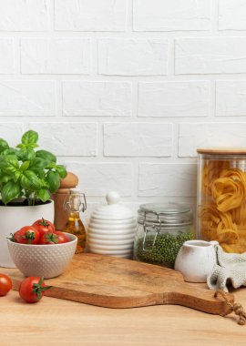 Boş ahşap kesme tahtası mutfak tezgahında oturur. Taze malzemeler, baharatlar ve çatal bıçaklarla çevrili. Kaba bir mutfak sahnesi yaratır.