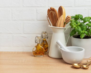 Mutfak tezgahı sahnesinde ahşap kaplar, fesleğen fabrikası, yağ şişeleri, havan ve tokmak var. Sıcak bir mutfak atmosferi yaratıyor.