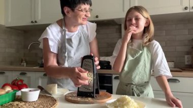 Büyük anne peyniri rendeliyor, torun ise parlak bir mutfakta tadını alıyor.