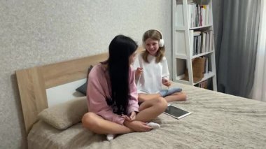 İki kız kardeş bir yatakta oturmuş, kulaklıklarını ve tabletlerini paylaşıyorlar.
