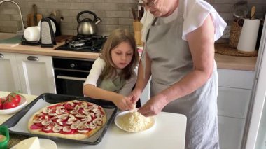 Büyükanne ve torun pizza hazırlıyor, peynir ekliyorlar.