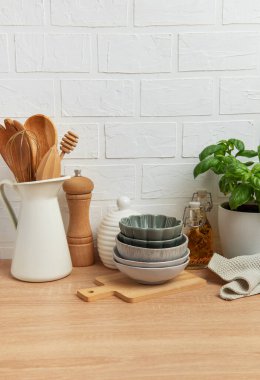 Kâseler, tahta kaşıklar, bir biber değirmeni, yağ şişeleri ve fesleğen bitkisi de dahil olmak üzere çeşitli mutfak malzemeleri beyaz tuğla bir duvara karşı ahşap bir tezgahta güzelce düzenlenmiştir.