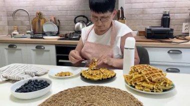 Evcil bir mutfakta krema ve yabanmersinli waffle kahvaltısı hazırlayan yaşlı bir kadın.