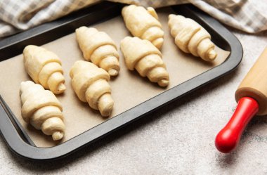 Fırın tepsisine yerleştirilmiş taze pişmemiş kruvasanlar, merdane ve mutfak havlusuyla birlikte fırına hazır.