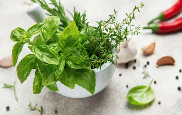 Taze fesleğen, kekik ve beyaz seramik bir havanda biberiye kabuklu, sarımsaklı karanfil, biberli ve gri arka planda kırmızı biberli.
