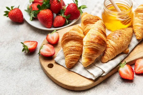 Taze pişmiş kruvasanlar lezzetli bir kahvaltı için ahşap bir tahtada çilek ve bal ile servis edilir.