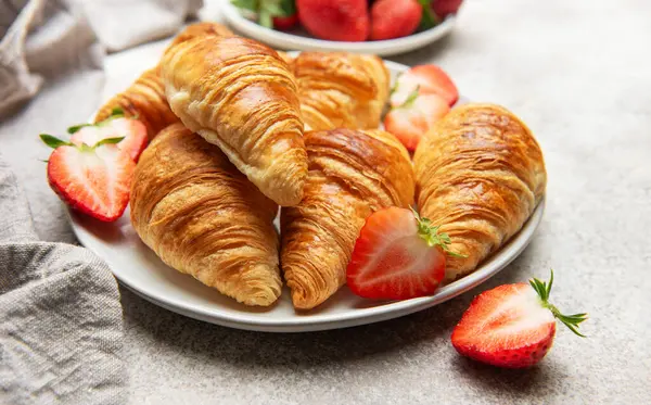Altın sarısı kruvasanlar, beyaz bir tabakta taze çilek ile güzel bir şekilde düzenlenir ve cazip bir kahvaltı sahnesi yaratır.