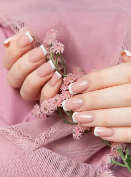 Fransız manikürünü gösteren bir kadın pembe arka planda küçük pembe çiçekler tutuyor.