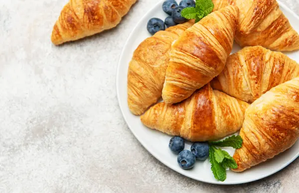 Lezzetli altın sarısı kruvasanlar taze yaban mersini ve nane yapraklarıyla beyaz bir tabakta servis edilir. Bu da cezbedici bir kahvaltı sahnesi yaratır.