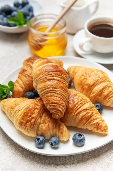 Taze pişmiş kruvasanlar beyaz bir tabakta yabanmersini, bal ve bir fincan kahveyle servis edilir, cezbedici bir kahvaltı sahnesi yaratır.