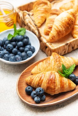 Taze pişmiş kruvasan, yaban mersini, nane yaprağı ve bal ile servis edilir ahşap tabakta lezzetli bir kahvaltı sahnesi yaratır.