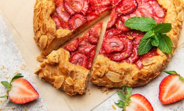 Badem yaprakları ve nane yapraklarıyla süslenmiş taze fırınlanmış çilek galeti, yakınlardaki taze çileklerle parşömen kağıdında dinleniyor.