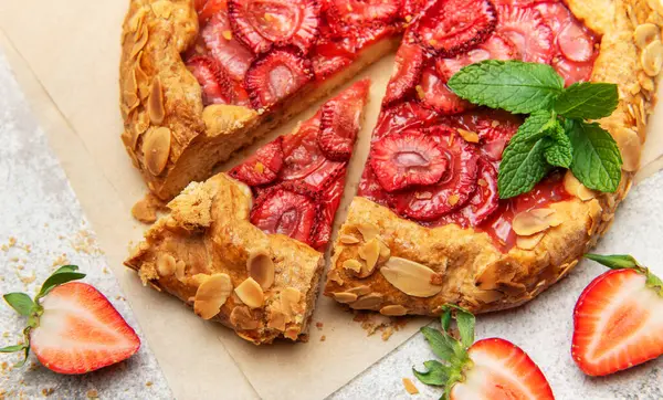 Badem yaprakları ve nane yapraklarıyla süslenmiş taze fırınlanmış çilek galeti, yakınlardaki taze çileklerle parşömen kağıdında dinleniyor.