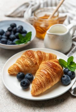 İki altın sarısı kruvasan taze yaban mersini, nane yaprağı, bal ve süt ile beyaz bir tabakta servis edilir ve nefis bir kahvaltı sahnesi yaratır.