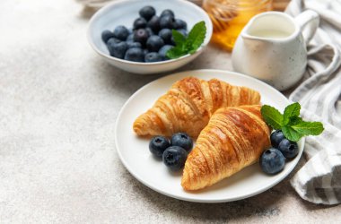 İki altın sarısı kruvasan taze yaban mersini, nane yaprağı, bal ve süt ile beyaz bir tabakta servis edilir ve nefis bir kahvaltı sahnesi yaratır.