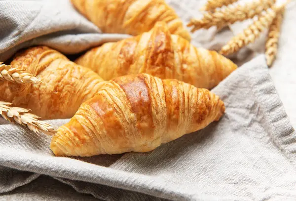 Altın sarısı kruvasanlar ve buğday sapları keten kumaş üzerinde samimi bir kahvaltı sahnesi yaratır.