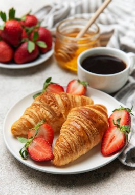 Taze pişmiş kruvasanlar beyaz bir tabakta çilek, bal ve kahveyle servis edilir ve nefis bir kahvaltı sahnesi yaratır.