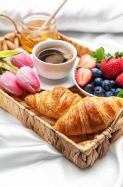 Kahve fincanı, kruvasan, taze böğürtlen, bal ve pembe lalelerle nefis kahvaltı tepsisi beyaz çarşaflar üzerinde yatakta servis edilir.