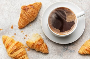 Altın rengi kruvasan ve dumanı tüten bir fincan kahve dokulu bir yüzey üzerinde cezbedici bir kahvaltı sahnesi yaratır.