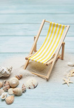 Minyatür iskemle deniz kabuklarıyla çevrili kumlu bir sahilde oturuyor. Yaz tatili ve rahatlama hissi uyandırıyor.