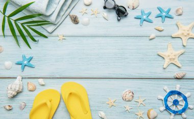 Yaz tatili arka planında parmak arası terlikler, havlu, güneş gözlüğü, deniz kabuğu ve açık mavi ahşap üzerinde yıldız balıkları yer alıyor.