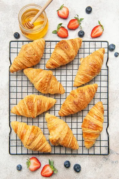 Lezzetli altın rengi kruvasanlar tel askıda soğuyor, yanında taze çilek, yabanmersini ve bir kavanoz bal var.
