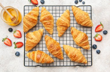 Lezzetli altın rengi kruvasanlar tel askıda soğuyor, yanında taze çilek, yabanmersini ve bir kavanoz bal var.