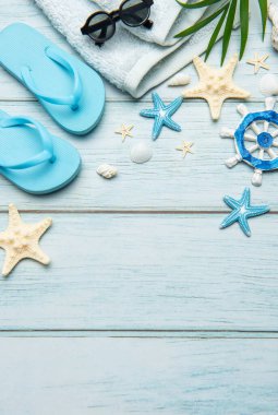 Terlikler, güneş gözlükleri, havlu, deniz kabukları, denizyıldızı ve gemi tekerleği açık mavi ahşap arka planda fotokopi alanı olan bir çerçeve oluşturuyor.