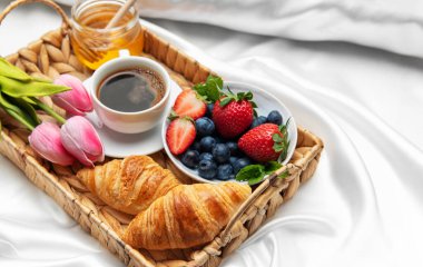 Kahve fincanı, kruvasan, taze böğürtlen, bal ve pembe lalelerle nefis kahvaltı tepsisi beyaz çarşaflar üzerinde yatakta servis edilir.