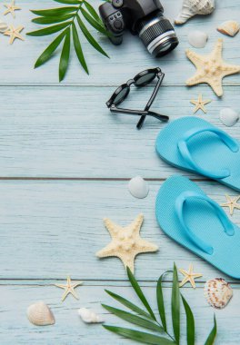 Parmak arası terlikler, kamera, güneş gözlüğü, deniz kabuğu ve palmiye yaprakları mavi ahşap bir arka planda yaz tatili düzenlemesi yapıyor.