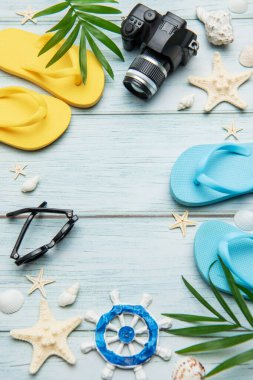 Mavi ahşap arka planda parmak arası terlikler, kamera, güneş gözlüğü, deniz kabuğu, deniz yıldızı ve palmiye yaprakları içeren yaz tatili rahatlama konsepti